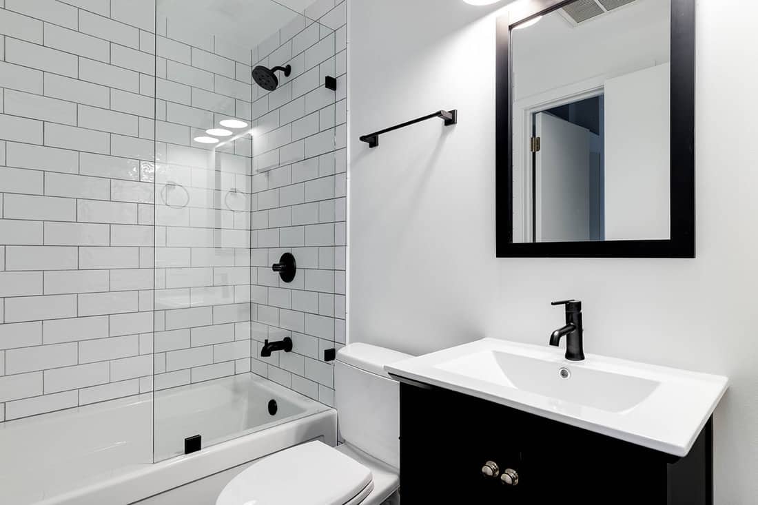 A small modern bathroom with a dark vanity, mirror frame, and hardware. White subway tiles line the bathtub and shower with black faucets