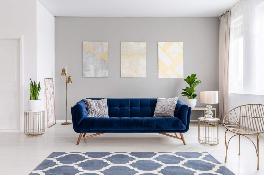 An elegant navy blue sofa in the middle of a bright living room interior with gold metal side tables and three paintings on a gray wall. 