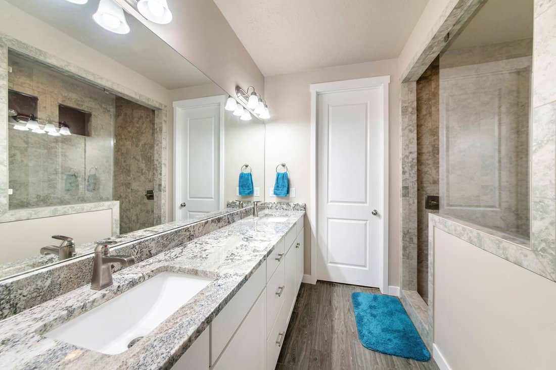 Bathroom with long vanity with two sinks and walk-in shower with half wall and glass. There is a mirror wall above the tile counter with towel hanging on side and vinyl wooden flooring with blue rug.