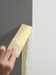 Hand taking off masking tape from the wall after painting. Home renovation tricks and minimalistic style., How Long After Painting To Remove Tape [And How To Remove It]