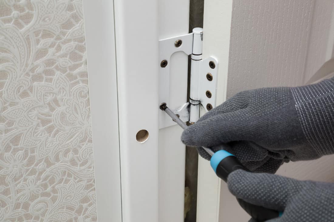 Man hands in protective gloves using manual screwdriver and screwing hinges on frame of wooden door to wall in bathroom. 