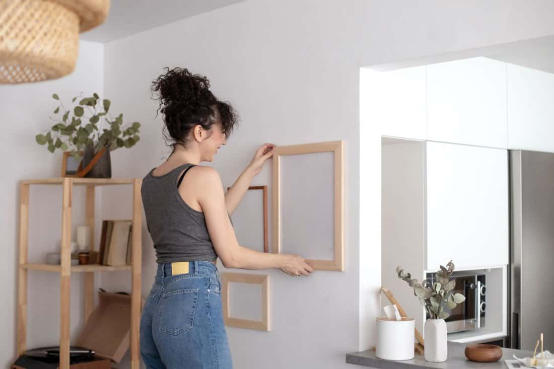 Modern female hanging wall pictures photos in wooden frame decorated scandi minimalistic style room apartment. Brunette curly woman attaching elegant decor elements cosiness home white light interior