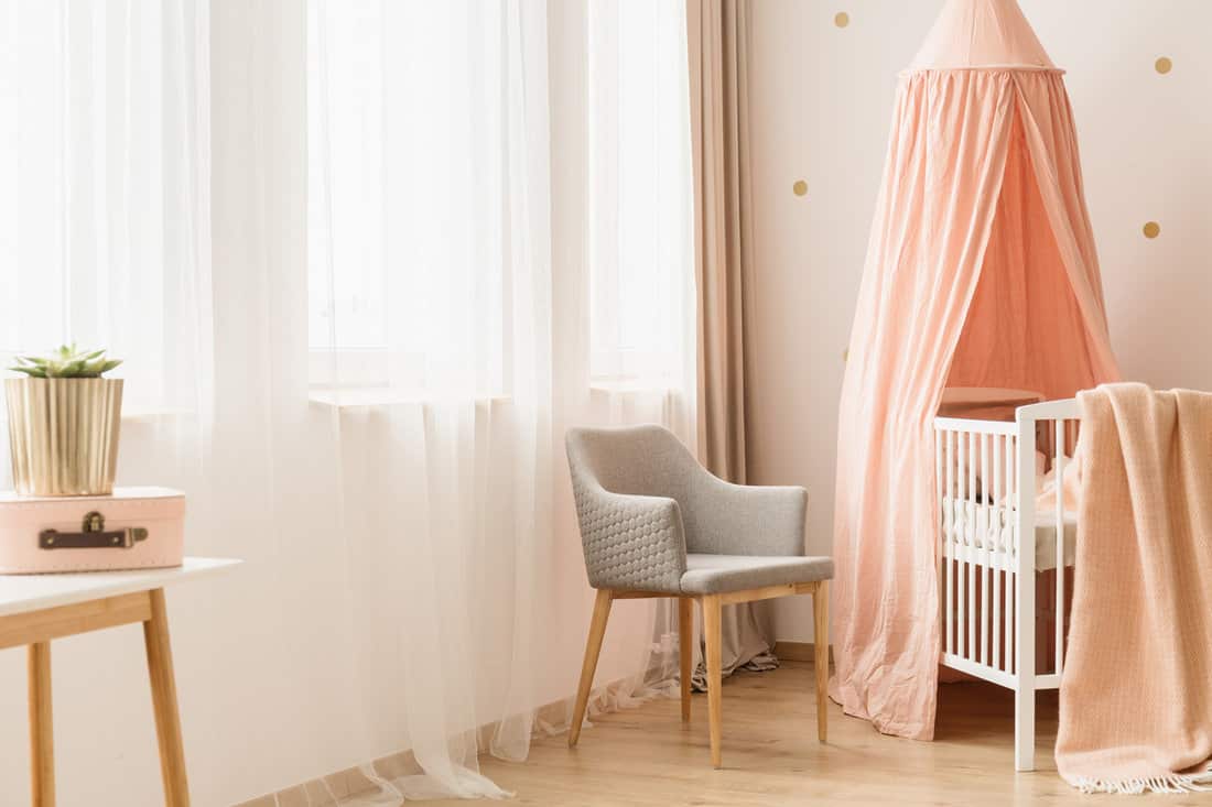 Modern, gray chair by windows and a cozy baby crib with peach pink canopy in a cute, bright nursery room interior