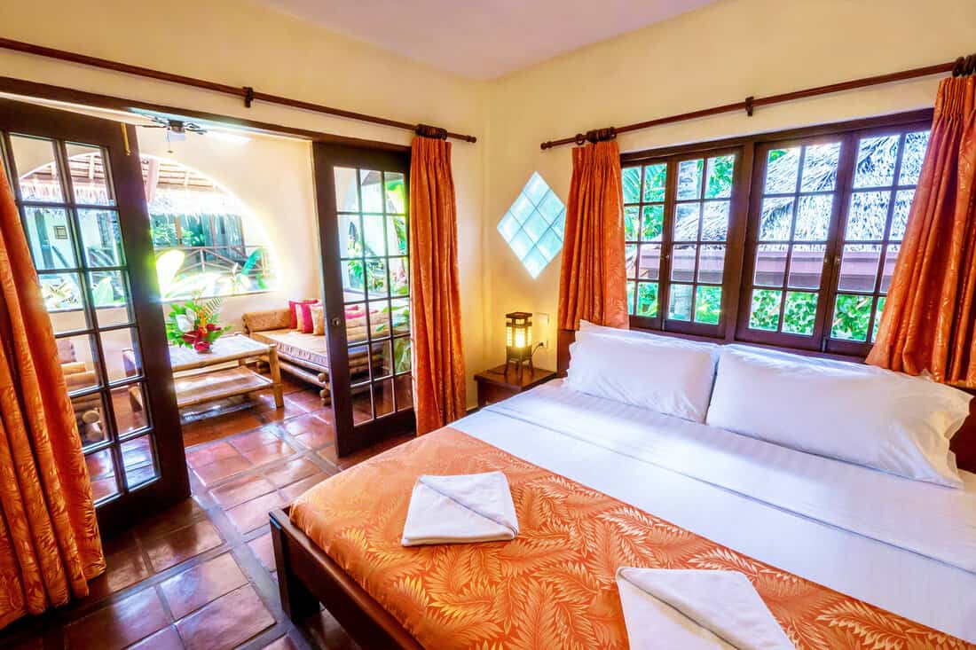 Pretty tropical resort bedroom with yellow walls and wooden french windows and doors opening onto a terra-cotta tiled balcony with rustic furniture, in a garden setting.