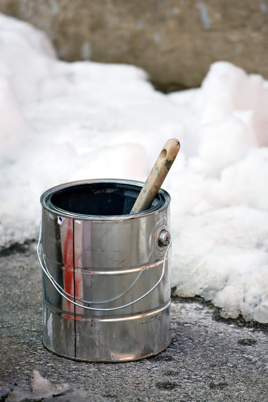 Paint can and brush on the ground outside in the winter. Colder temperatures call for specialty paints made with different formulas.