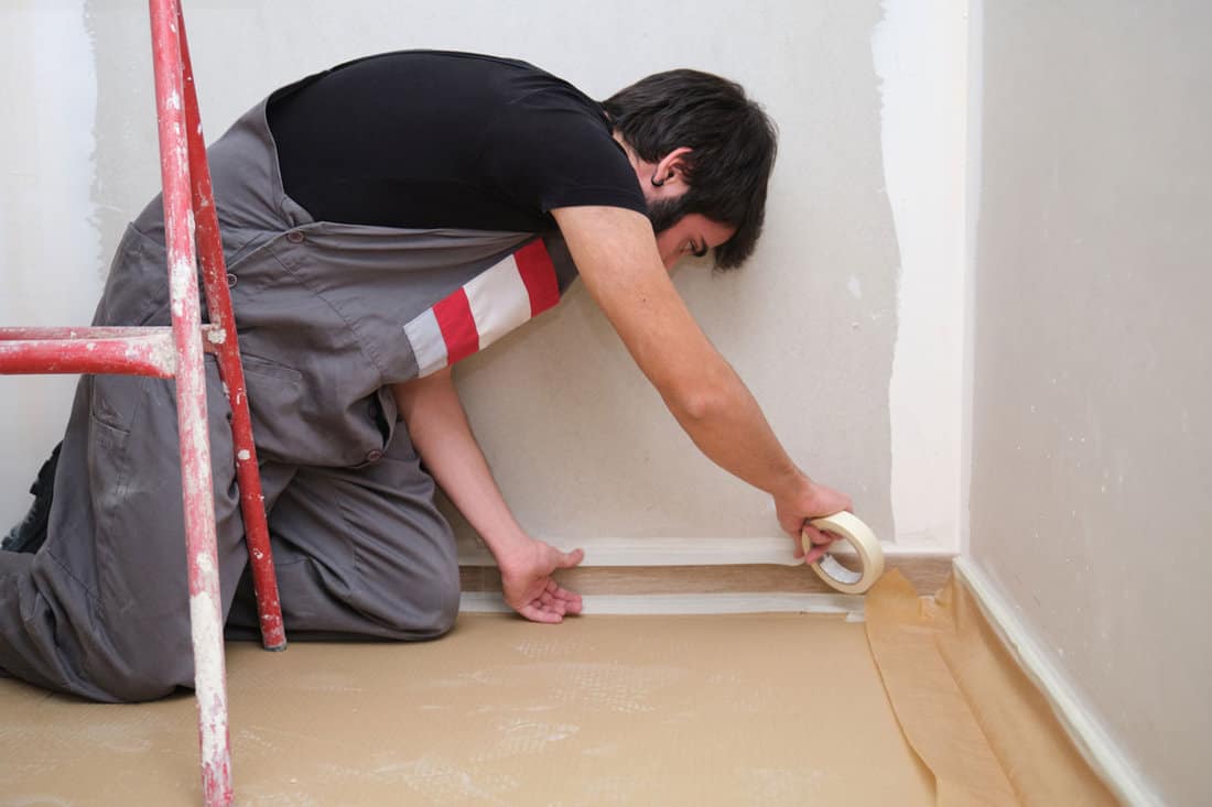 Painter using tape to cover the skirting board before painting the room