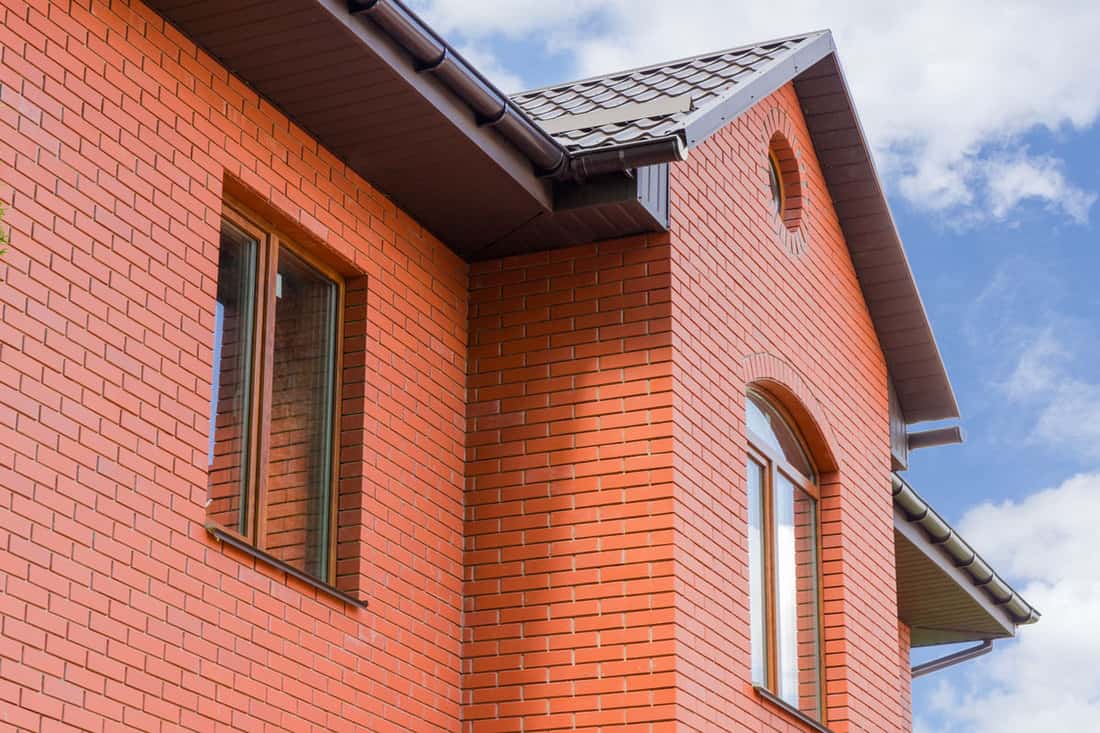 Part of wall made of the red brick, windows and fragment of roof of modern private dwelling house against of sky