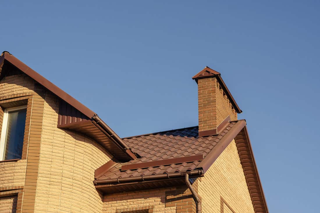 The brown roof with mantel chimney of a beautiful modern stone mansion against a cloudless sky. Brown roof with gable, gutter system and snow guards. Contemporary real estate, architecture.