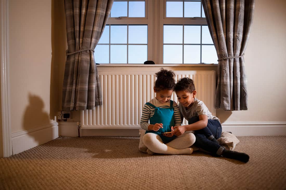 Two siblings playing on a mobile phone together while sitting in a room on the floor.