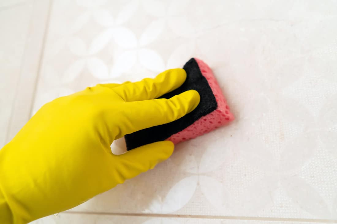 A hand in a yellow glove holds a sponge and wipes the tiles on the wall in the bathroom, a man does cleaning with detergent at home. 
