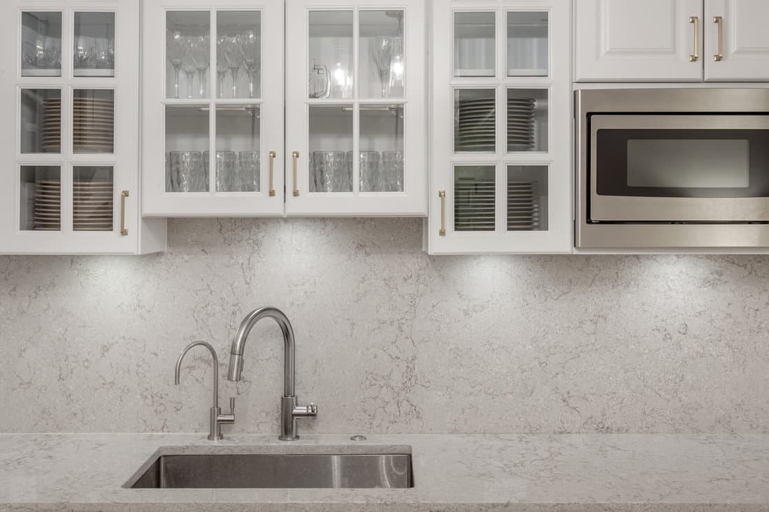 Glass door white kitchen cabinets with stainless steel microwave, kitchen faucet, soap dispenser, avonite countertop and avonite backsplash with gold color handles