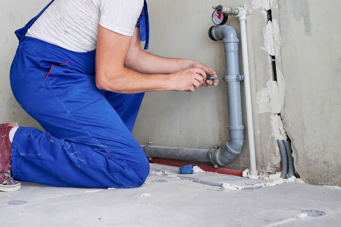 Horizontal snapshot of young plumber working with grey sewer pipes, fixing them to wall