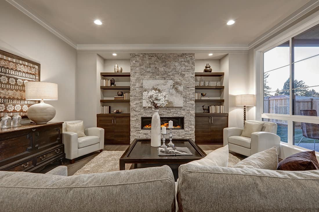 Living room interior in gray and brown colors features stone fireplace with built-in shelves and cabinets, large dark stained cocktail table atop beige fluffy rug and large window