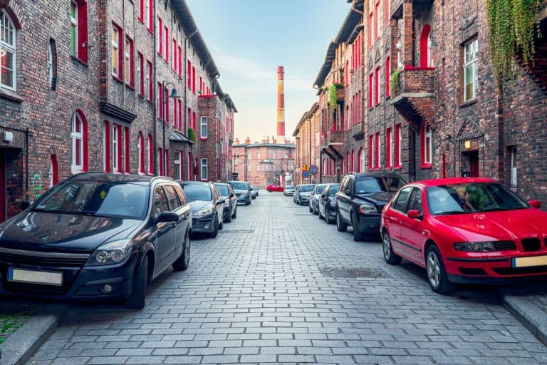 Renovated, old, brick block houses, cobblestone street, cars parked on the street, factory chimney and car on side, What Is Considered 'Blocking' A Driveway Or Sidewalk?