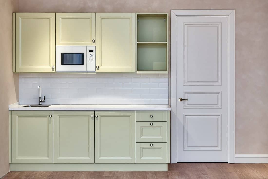 Small kitchen furniture set in classic decorative style with sink and microwave oven near white door in light spacious room