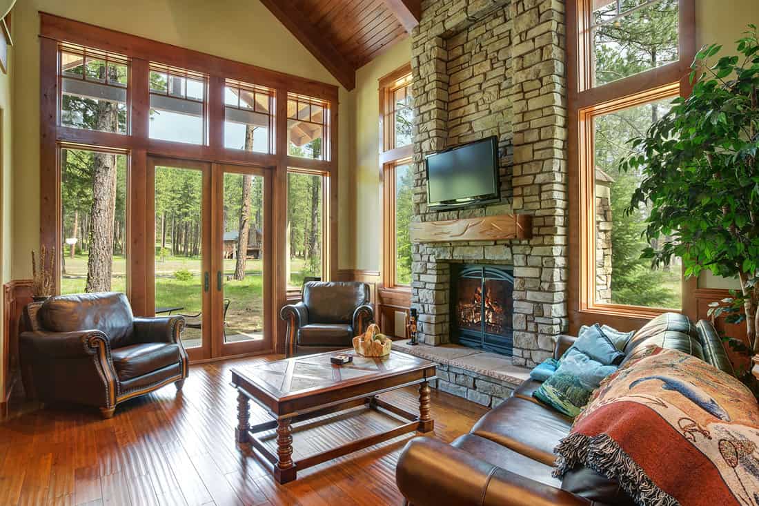 Stunning living room design includes a stone wall fireplace, brown leather sofa with a curved cocktail table facing a pair of leather armchairs with nail head trim atop hardwood floor