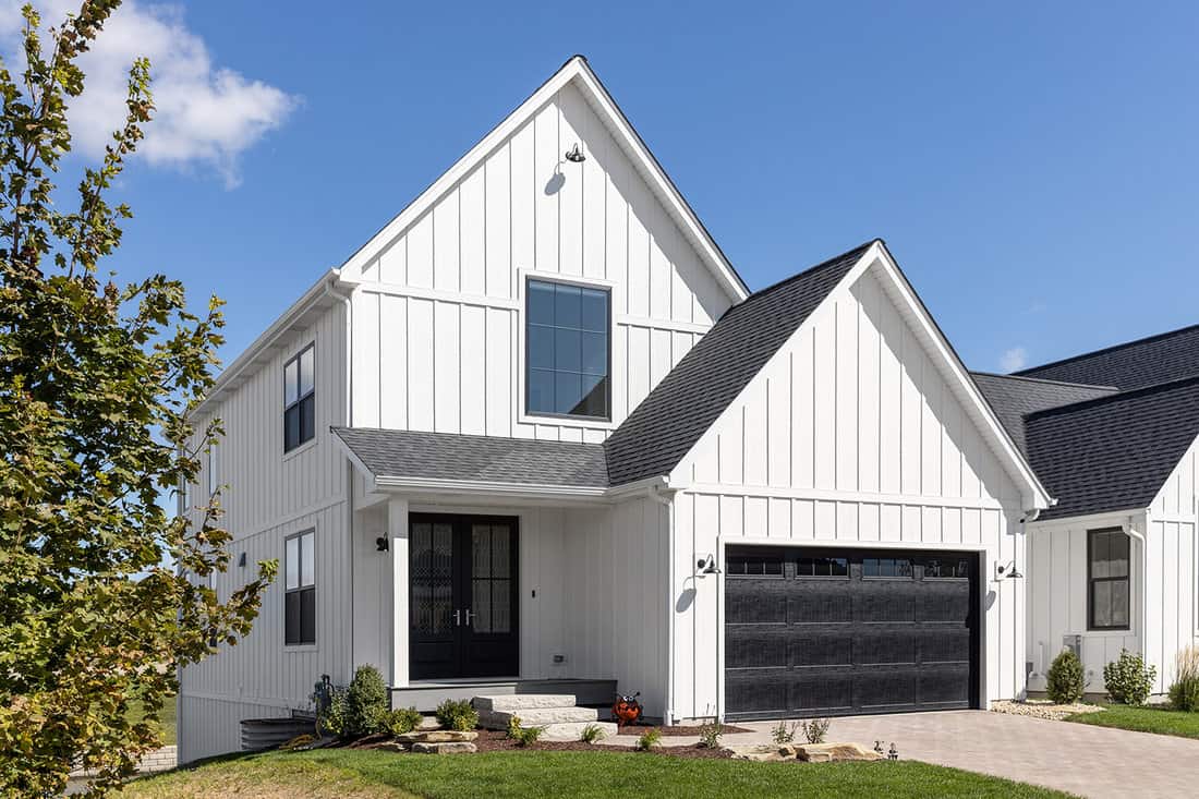 A large, white modern farmhouse with vertical siding, a black two car garage, and lights mounted on the siding