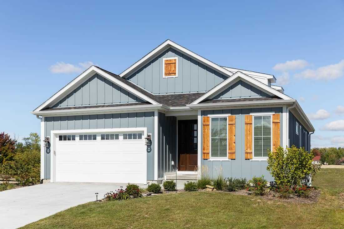 A light blue ranch home with a two car garage, wood window shutters, vertical siding, and a dark wood front door