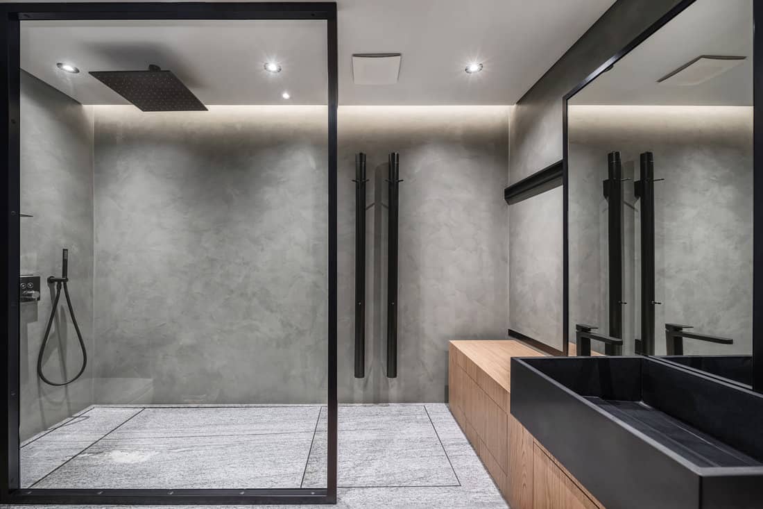 Bathroom in a modern style with gray tiled walls. There is a shower with a glass partition, wooden stand with a black sink and a faucet, large mirror, luminous lamps. Horizontal. 