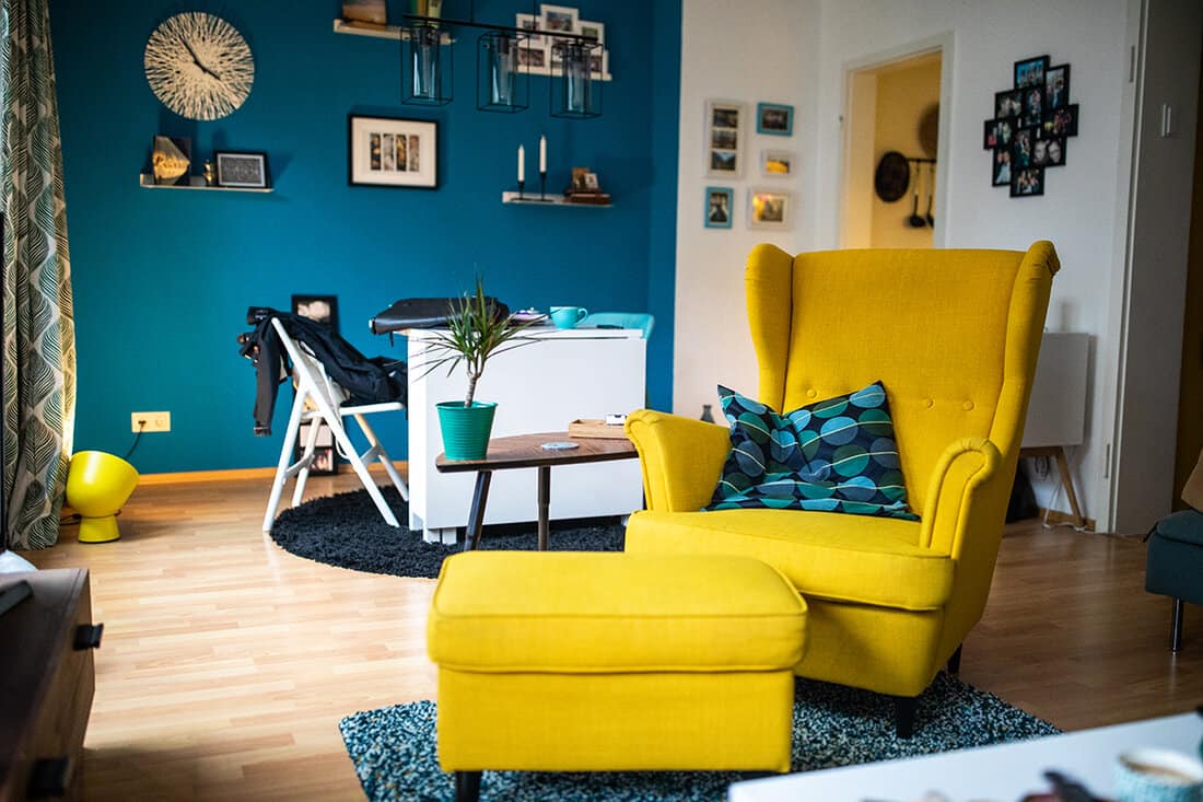 Bright living room with yellow armchair, blue rug, blue pillow, table, potted plant, desk and parquet floor