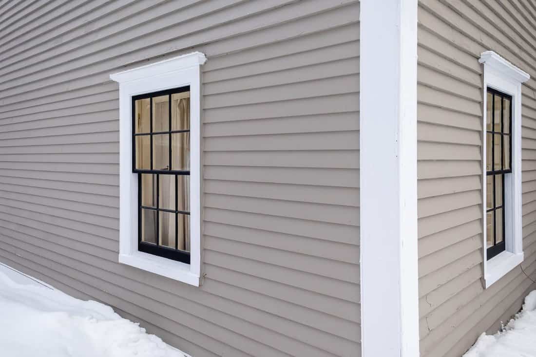 Corner view of a vintage tan coloured house with white trim and black wooden window spacers. The glass in the window is wavy and reflecting the sun. There's snow on the ground and up the building. 