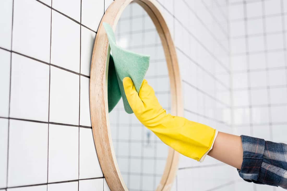 Cropped view of woman in rubber glove swiping mirror with rag while cleaning bathroom