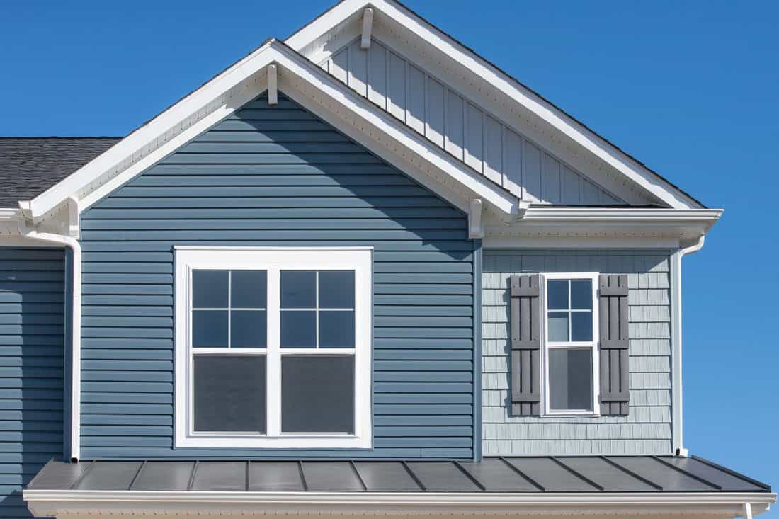 Elegant blue horizontal vinyl siding, shingle and vertical siding in a double gable roof with white decorative corbels, double sash window with matching shut