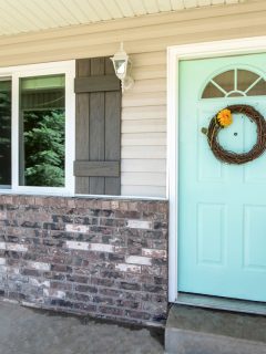 Entrance of a house with brick walls and vinyl sidings. Front door exterior with a mint green door with arched glass panel and leafless wreath beside the double hung window., Should I Paint Brick And Siding The Same Color?