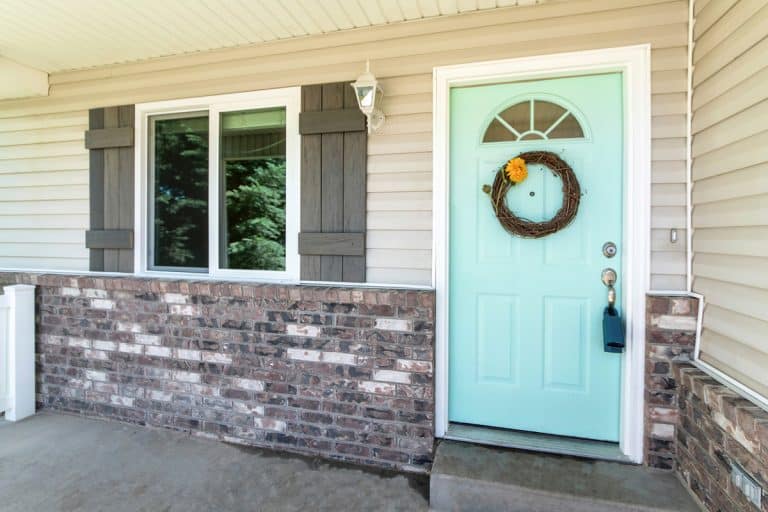 Entrance of a house with brick walls and vinyl sidings. Front door exterior with a mint green door with arched glass panel and leafless wreath beside the double hung window., Should I Paint Brick And Siding The Same Color?