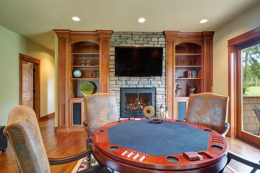 Fun game area on the second floor. Poker table surrounded by chic gray leather chairs facing stone wall fireplace flanked by built in bookshelves.