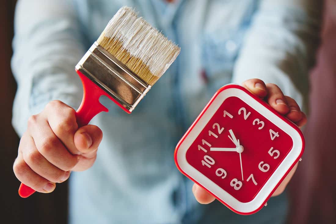 Men's hands hold the brush with white paint and analogue clock.