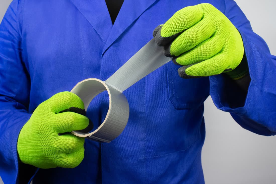 Photo of a worker in blue outfit and green gloves holding duct tape. 