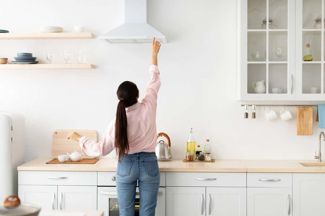 Rear back view of young millennial housewife choosing and selecting mode on range exhaust hood, pushing button on mechanical fan above the stove
