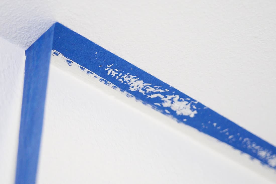 Removing masking tape from ceiling. A painter pulls of blue painter's tape from the wall to reveal a clean edge with the ceiling. 