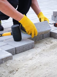 The master in yellow gloves lays paving stones in layers. Garden brick pathway paving by professional paver worker. Laying gray concrete paving slabs in house courtyard on sand foundation base., Can You Cement Over Pavers?