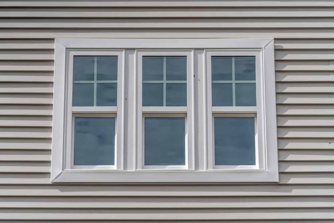 Triple hung window with fixed top sash and bottom sash that slides up, sash divided by white grilles a surrounded by white elegant frame horizontal white vinyl siding