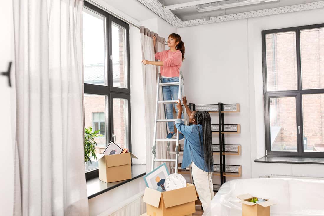 moving, home improvement and real estate concept - happy smiling women with ladder hanging curtain 