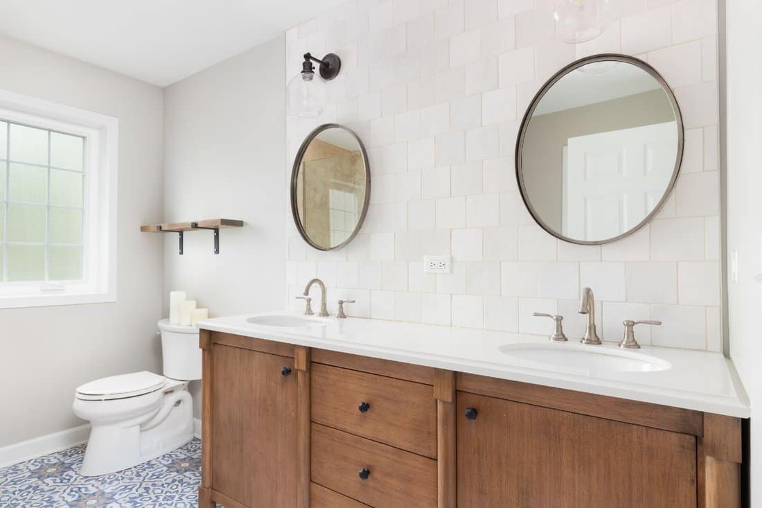 A cozy bathroom with a patterned tile floor, natural wood vanity, tiled backsplash, and lights mounted above circular mirrors