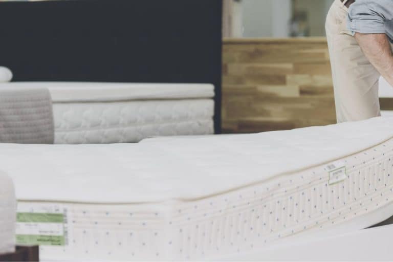 A man in a gray shirt and white pants chooses a mattress in a large magah of beds, mattresses and bed linen. He is just examining one of them. - How To Tell What Sleep Number Bed You Have