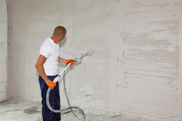 A man is applying stucco on the wall, plastering, coating the wall by spraying stucco with a stucco sprayer., At What Temperature Can You Apply Stucco?