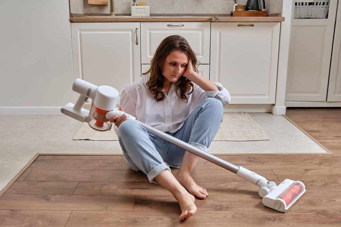 A sad woman with a wireless portable vacuum cleaner in the kitchen. A tired woman cleans the floor in an apartment with a vacuum cleaner