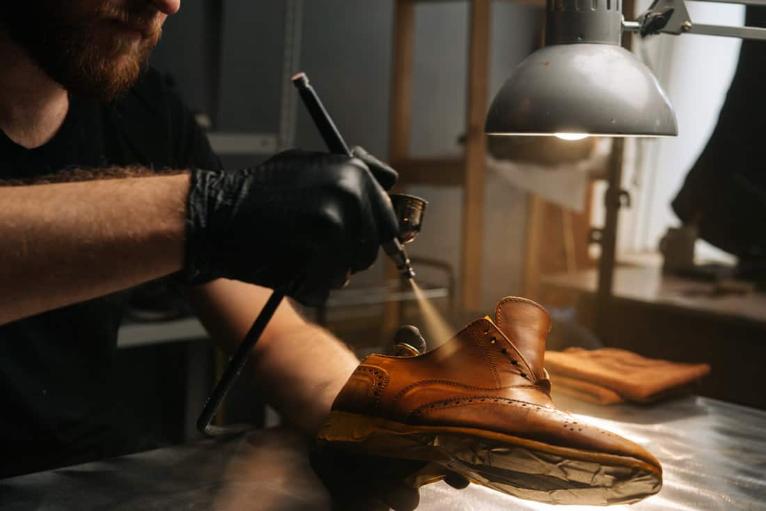 Close-up hands of unrecognizable shoemaker wearing black gloves spraying paint of light brown leather shoes.