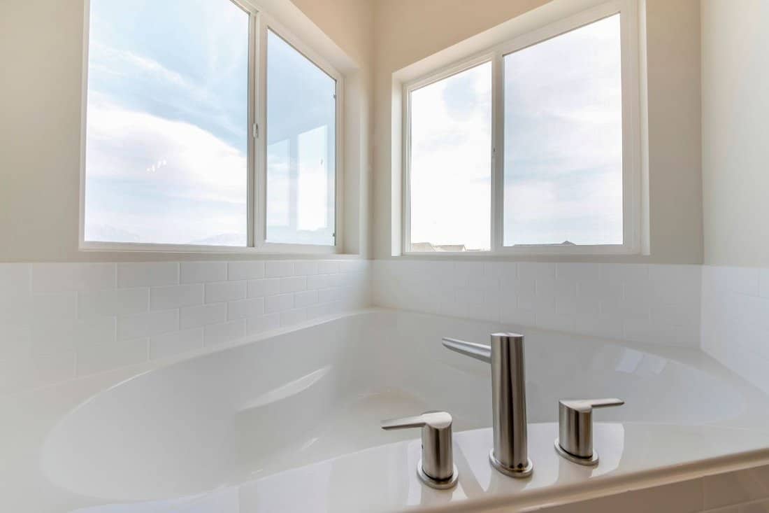 A corner bathtub in a bathroom with widespread faucet against the windows