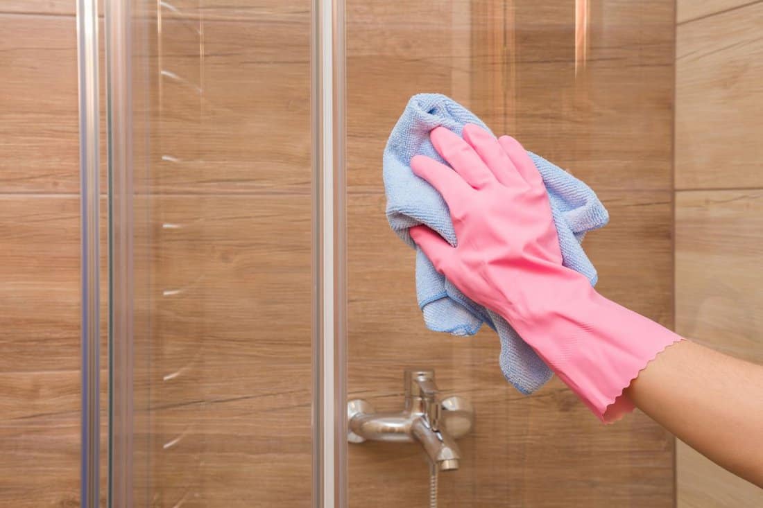 Employee hand in rubber protective glove with rag washing and polishing a glass shower doors. Housewife cares about house. Spring general or regular clean up. Commercial cleaning company concept.