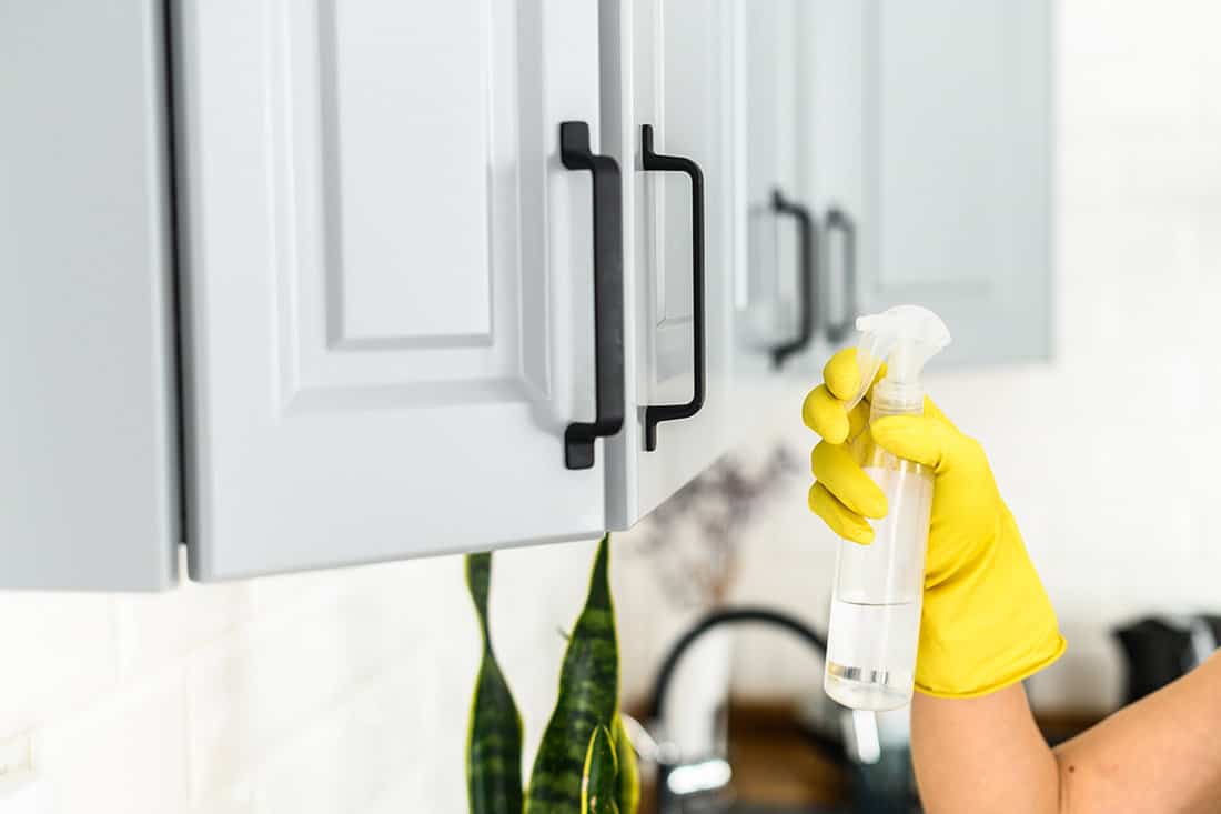 Hands in yellow protective rubber gloves is cleaning handles of kitchen cabinets at home