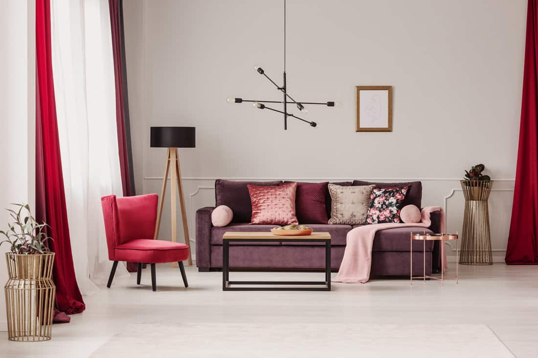 Lamp above table in sophisticated living room interior with red armchair and violet sofa against the wall with a poster