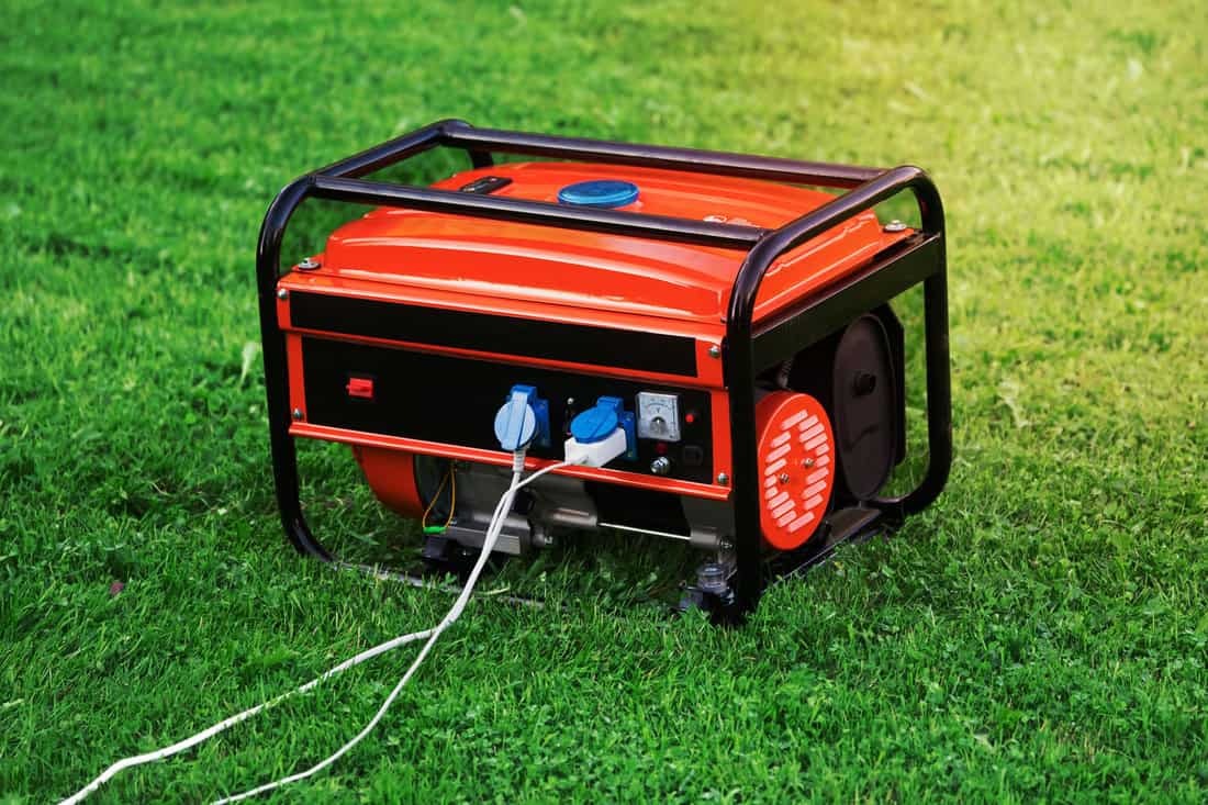 Portable electric generator with white electric wires connected to it on the green grass in summer day outdoors. The generator provides with electricity electrical appliances in nature 