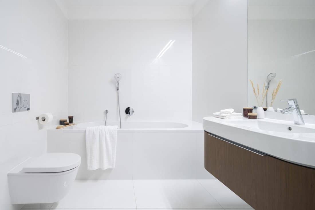 Spacious and elegant bathroom with big bathtub, white tiles, washbasin with wooden cabinet with drawer and big mirror
