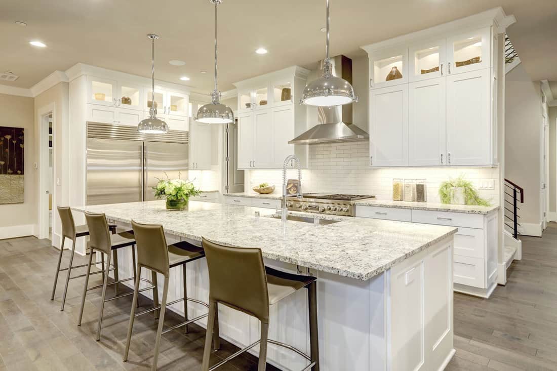 White kitchen design features large bar style kitchen island with granite countertop illuminated by modern pendant lights. Stainless steel appliances framed by white shaker cabinets .