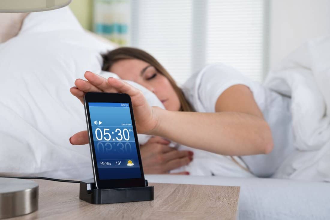 Woman Lying On Bed Snoozing Alarm On Mobile Phone Screen